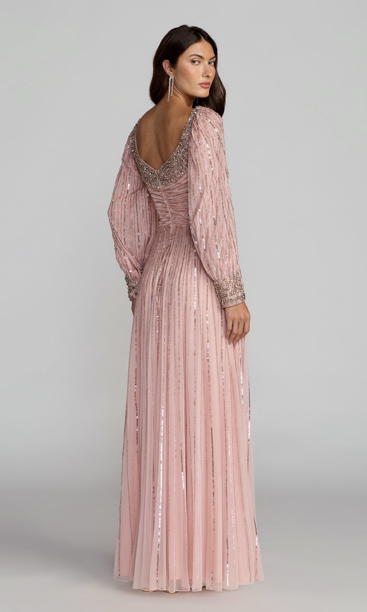 A woman stands against a plain background, looking over her shoulder and wearing the Long Formal Dress: Mac Duggal 23085—a blush pink gown with sequins, long sleeves, and a scoop back that exudes timeless elegance and sophistication.
